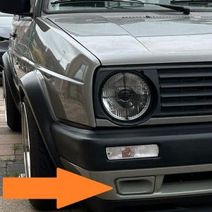 Könnte beinhalten: Die Frontpartie eines grauen Autos mit rundem Scheinwerfer, schwarzem Kühlergrill, rechteckigem Blinker und Lufteinlass im unteren Stoßfänger. Ein großer orangefarbener Pfeil zeigt auf den rechteckigen Lufteinlass im vorderen Stoßfänger.