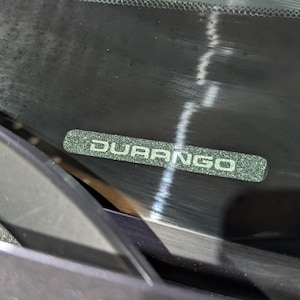 Puede incluir: Primer plano del parabrisas de un vehículo con la palabra "DURANGO" en un emblema rectangular texturizado verde plateado. El parabrisas es oscuro y reflectante, con un limpiaparabrisas visible en la parte inferior.