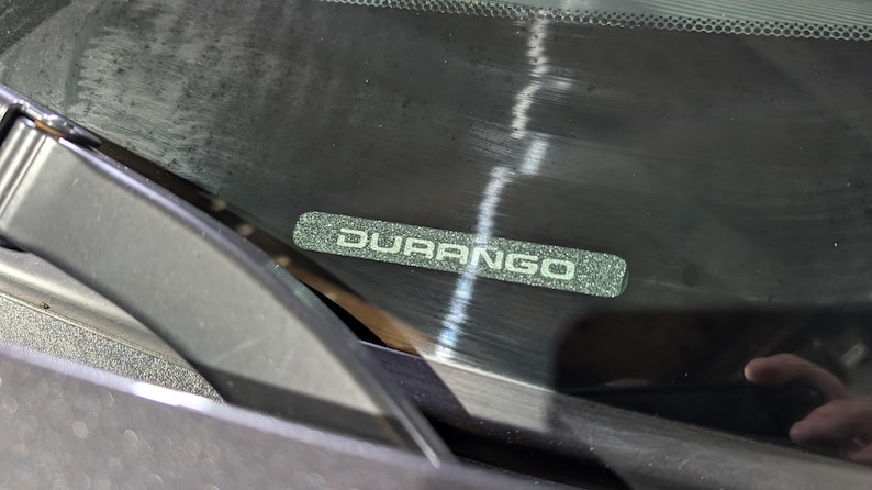 Op de afbeelding: Close-up van de voorruit van een voertuig met het woord "DURANGO" in een rechthoekig, getextureerd, zilvergroen embleem. De voorruit is donker en reflecterend.