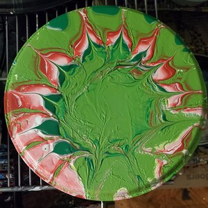 May include: A round, green, red, and white painted plate with a swirling, marbled design. The design resembles a flower with petals.