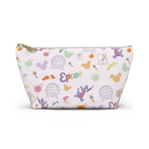 Peut inclure: Trousse de maquillage blanche avec fermeture éclair dorée, ornée d'un motif coloré de taches de peinture, de pinceaux, de silhouettes de Mickey Mouse et du mot "Epcot". Le motif comprend un dragon violet et une sphère géodésique.