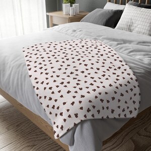 May include: A white blanket featuring a repeating pattern of dark brown floral shapes. The blanket is draped over a bed with white sheets and pillows. The bed is made of light-coloured wood.