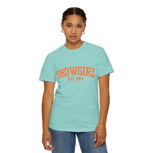 May include: A light turquoise short-sleeved t-shirt featuring the text "SHOWGIRL" in a bold, arched orange font, with the text "EST. 2025" printed directly underneath in a smaller, matching orange font across the center of the chest.