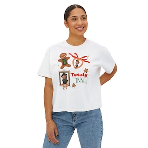 May include: White t-shirt with a festive graphic. The design includes a gingerbread man, a heart-shaped ornament with a ribbon, and a framed elf, all with the text "Totally Tinsel". The shirt is worn by a person.