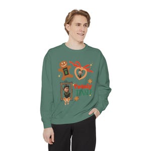 May include: Green sweatshirt with a festive design. The graphic includes a gingerbread man, a heart with a character, and a framed portrait. The text "Totally Tinsel" is also featured. The sweatshirt is a comfortable, casual style.