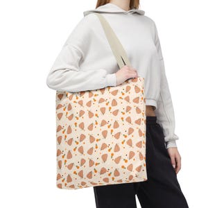 Peut inclure: Un sac fourre-tout avec un motif répétitif de citrouilles orange et de petites fleurs orange sur un fond crème. Le sac possède une large bandoulière de couleur claire et une forme rectangulaire, conçue pour transporter des objets facilement.
