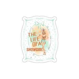 May include: A die-cut sticker featuring a light green scalloped border with a polka dot pattern. Inside an oval frame, a showgirl illustration stands next to the orange text THE LIFE OF A SHOWGIRL, accented by a light blue bow and star sparkles.