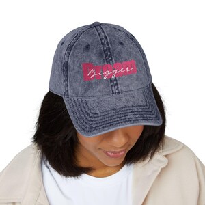 May include: A denim blue baseball cap with the word "Bigger" in pink script. The cap has a curved brim and a distressed, vintage look. The hat is a casual accessory.