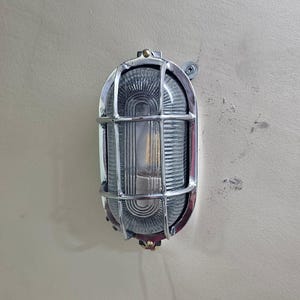 Op de afbeelding: Een ovaalvormige, verchroomde wandlamp. De lamp heeft een geribbelde glazen lens, beschermd door een metalen kooi. De lamp is bevestigd aan een beige muur. Een schroef is zichtbaar.