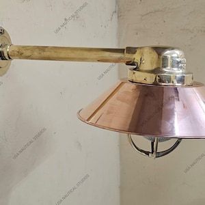 Könnte beinhalten: Eine nautische Wandleuchte aus Messing mit einem Kupferschatten. Die Leuchte hat einen langen Arm und einen runden Sockel. Der Schirm ist nach unten geneigt.