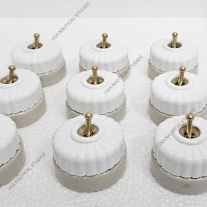 Op de afbeelding: Een verzameling witte keramische lichtschakelaars met messing tuimelschakelaars. De schakelaars hebben een geribbeld ontwerp en een grijze basis. De afbeelding bevat de tekst "USA NAUTICAL STUDIOS".