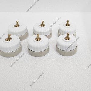 Könnte beinhalten: Sechs weiße, keramische Lichtschalterabdeckungen mit goldenen Kippschaltern. Die Abdeckungen haben ein dekoratives geriffeltes Design und eine beige Basis.