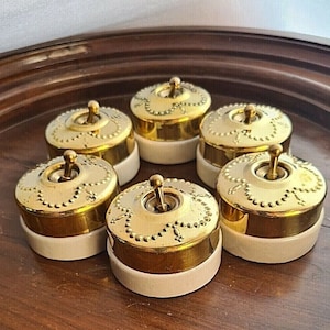 May include: Six vintage-style light switches with gold-colored metal accents and a cream-colored base. The switches have a decorative design around the toggle and are arranged on a wooden surface. The switches are a decorative element.