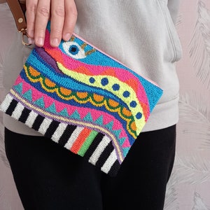 May include: A colourful beaded clutch with a whimsical design featuring a large eye, geometric patterns, and stripes. The clutch has a black and white striped bottom and a pink zipper.