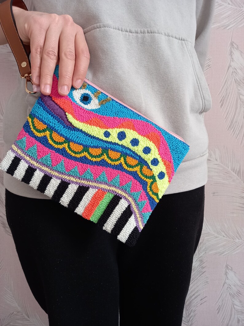 May include: A colourful beaded clutch with a whimsical design featuring a large eye, geometric patterns, and stripes. The clutch has a black and white striped bottom and a pink zipper.