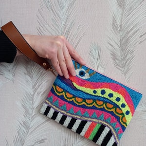 May include: A colourful beaded clutch with a pink lining and a brown strap. The clutch features a geometric design with a wavy pattern and a large eye in the centre.