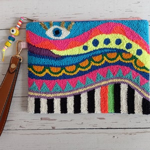 May include: A colourful, hand-stitched clutch bag with a bright blue eye design. The bag has a brown leather strap and is made with a variety of colourful yarns.