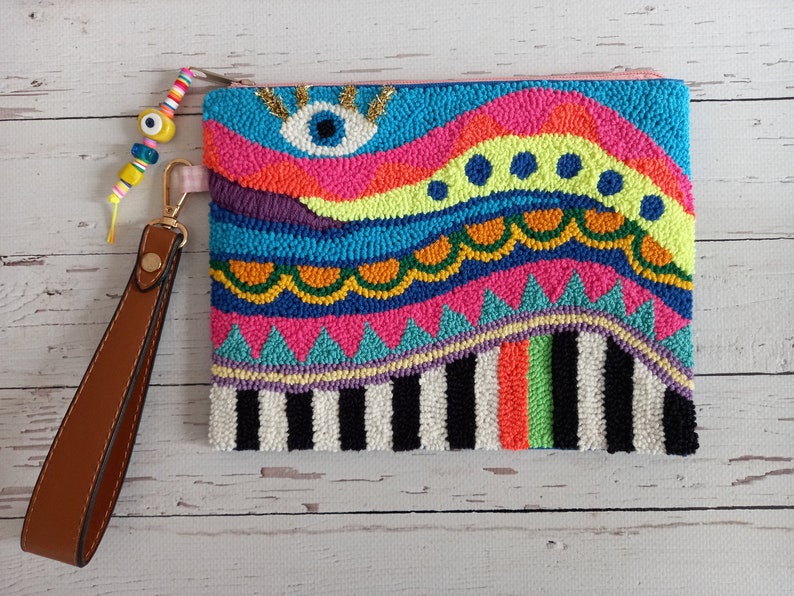 May include: A colourful, hand-stitched clutch bag with a bright blue eye design. The bag has a brown leather strap and is made with a variety of colourful yarns.