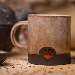 Puede incluir: Una taza de cerámica con un esmalte marrón y blanco. La taza tiene un pequeño agujero redondo en la parte inferior con un esmalte rojo y naranja en el interior. La taza está sobre una superficie de madera.