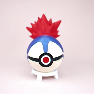 Puede incluir: Una Pok&eacute; Ball roja y azul con un dise&ntilde;o rojo y blanco. La Pok&eacute; Ball est&aacute; en un soporte blanco.