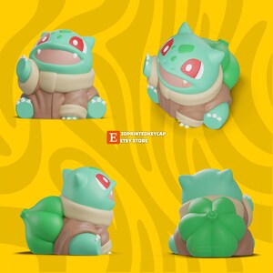 May include: A green and brown 3D printed keycap of a Pokemon character, Bulbasaur. The keycap is in a sitting position with its arms outstretched and its mouth open. The keycap is on a yellow background with a swirl pattern.