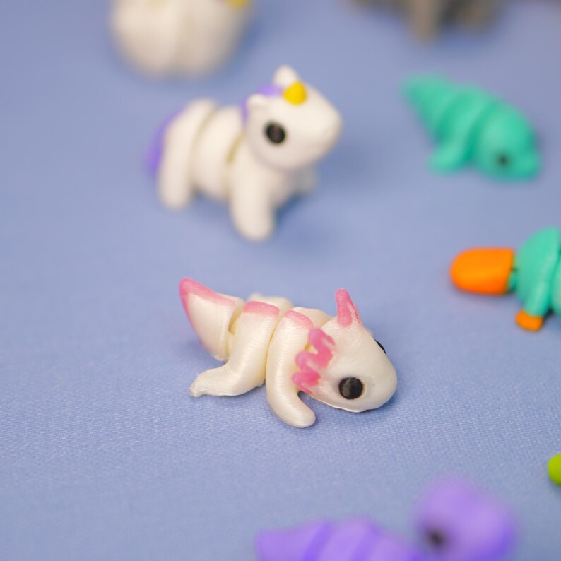 Op de afbeelding: Een verzameling kleine, witte, gearticuleerde dierenfiguren. De figuren omvatten een eenhoorn, een axolotl en een vogelbekdier. De figuren zijn gemaakt van glad, wit plastic en hebben roze accenten.