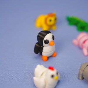 Op de afbeelding: Een set kleine, kleurrijke, 3D-geprinte dierenfiguren. De figuren omvatten een pingu&iuml;n, een leeuw, een krokodil, een kip en een worm. De figuren staan ​​allemaal op een lichtblauw oppervlak.