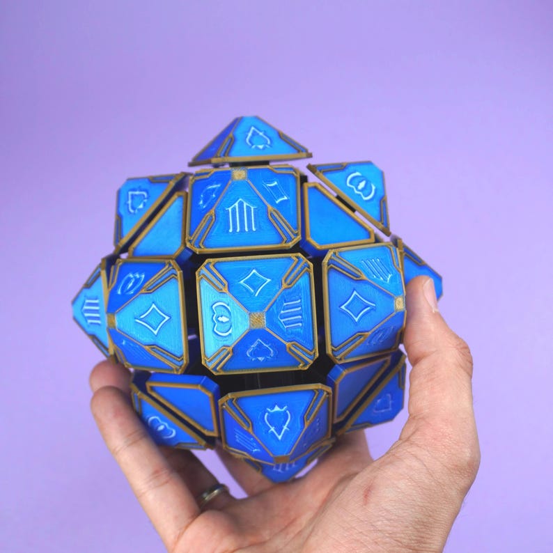 May include: A blue and gold geometric dice with various symbols on each face. The dice is being held in a hand.