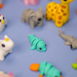 Op de afbeelding: Een verzameling kleine, kleurrijke, 3D-geprinte dierenfiguren. De figuren omvatten een dolfijn, een vogelbekdier, een eenhoorn, een koala, een kip, een giraffe en een haai.