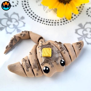 Könnte beinhalten: Eine braune und beigefarbene Tonskulptur einer Cartoon-Kreatur mit großen Augen und einem kleinen Quadrat aus gelbem Ton auf dem Kopf.