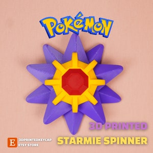 May include: A 3D-printed Starmie spinner in purple, yellow, and red. The spinner has a star-shaped design with a central red octagon and yellow accents. The Pokémon logo is visible above the spinner.
