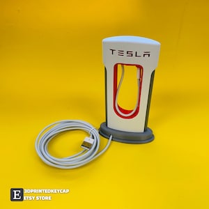 Puede incluir: Soporte de estación de carga Tesla impreso en 3D blanco y gris con un puerto de carga rojo y un cable blanco. El soporte tiene la palabra "TESLA" impresa en la parte delantera.