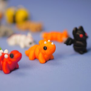 Op de afbeelding: Een verzameling kleine, handgeschilderde figuurtjes in verschillende kleuren, waaronder een rode draak, een oranje dinosaurus, een witte kat, een zwarte vleermuis en een gele bij. De figuurtjes zijn gemaakt van polymer clay en hebben een speelse, cartoonachtige stijl.
