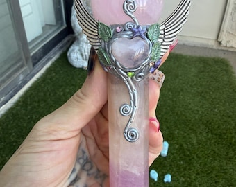 May include: A wand featuring a spherical rose quartz top and a hexagonal rose quartz base. The center is adorned with a silver-toned metal frame shaped like a heart, flanked by two wings, and decorated with small green leaves and purple accents.