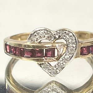 May include: A gold ring with a heart-shaped design set with small diamonds and red rubies. The heart is outlined with diamonds and the band is set with red rubies.