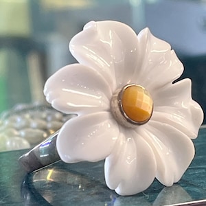 以下が含まれることがあります： 中央に白い彫刻が施された大きな花形のパーツがあしらわれた指輪。白い花の中央には、銀色の金属製のベゼルにセットされた、小さな丸いファセットカットの黄色い石が飾られている。リング部分は銀色の金属製である。