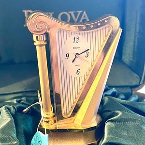 Może przedstawiać: Złoty zegar kwarcowy Bulova w kształcie harfy. Tarcza zegara jest biała z czarnymi cyframi i wskazówkami. Zegar jest prezentowany na zielonej satynowej powierzchni.