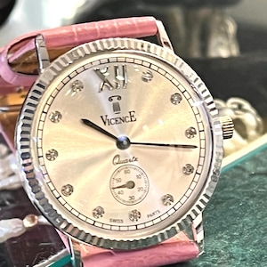 May include: A Vicence quartz watch with a silver-toned dial, a fluted bezel, and a pink textured leather strap. The dial features a Roman numeral at the twelve o'clock position, crystal hour markers, and a small sub-dial for seconds.