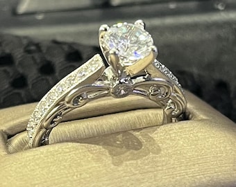 May include: A silver-toned ring featuring a large, round, brilliant-cut center stone held by four prongs. The band is decorated with intricate scrollwork and small, clear pavé-set stones along the shoulders, creating a detailed metal design.