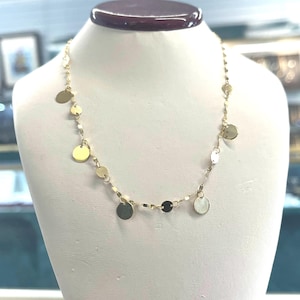 Può includere: Una collana d'oro con piccoli ciondoli circolari pendenti. La collana è esposta su un manichino bianco. I ciondoli sono di varie dimensioni e riflettono la luce. La collana è delicata ed elegante.