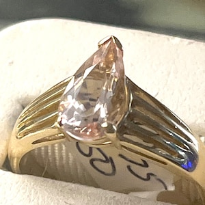 May include: A gold ring with a pear-shaped pink gemstone. The ring has a band with multiple grooves.