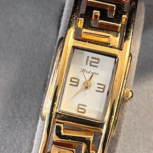 May include: A gold-tone wristwatch with a rectangular white face featuring the brand name Medana, gold-tone hands, and gold-tone numerals at 12, 3, 6, and 9. The watch band features a repeating geometric Greek key pattern in polished gold-tone metal.