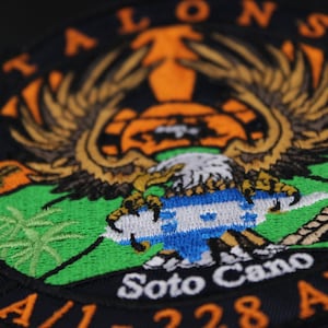 Puede incluir: Parche bordado con un diseño de águila con las alas extendidas, sobre un fondo verde y naranja. El parche incluye el texto "TALONS" y "Soto Cano A/1-228 AVN". El diseño incorpora una bandera.