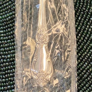 May include: A silver spoon in a clear plastic wrapper. The spoon has a decorative floral design on the handle and a rounded bowl. The spoon is likely part of a silverware set.