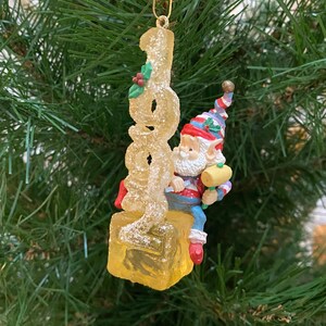 Könnte beinhalten: Weihnachtsbaumschmuck mit der Jahreszahl "2000" in einem klaren, glitzernden Design und einer kleinen Zwergenfigur, die einen Schlägel hält. Der Zwerg trägt einen bunten Hut und eine blaue Hose. Der Schmuck hängt an einem goldenen Haken.