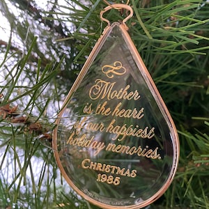 Könnte beinhalten: Transparenter Weihnachtsbaumschmuck aus Glas mit goldfarbenem Rand und goldfarbener Inschrift. Der Text lautet: „Mutter ist das Herz unserer schönsten Urlaubserinnerungen. Weihnachten 1985.“