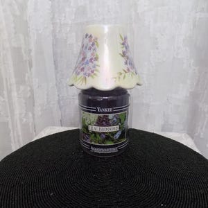 Pode incluir: Uma vela em frasco Yankee Candle Housewarmer roxo escuro com o rótulo Lilac Blossoms, coberta por uma cúpula de cerâmica creme com borda recortada e um padrão pintado de pequenas flores roxas e azuis com folhas verdes.
