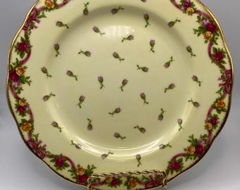 Könnte beinhalten: Ein cremefarbener Porzellanteller mit goldfarbenem Rand und einem Blumendesign. Der Teller hat einen Rand aus rosa Rosen, grünen Blättern und goldenen Bändern. Die Mitte des Tellers ist mit einem verstreuten Muster aus kleinen rosa Rosen verziert.