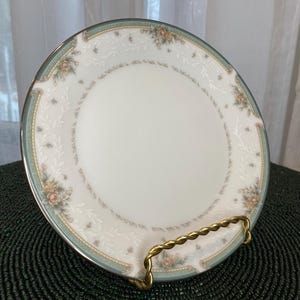 Puede incluir: Un plato de porcelana blanca con un borde floral verde y dorado. El plato está en un soporte dorado.