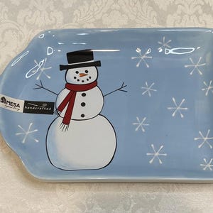 May include: A light blue rectangular serving tray with a snowman design. The snowman has a black top hat, a red scarf, and a carrot nose. White snowflakes are scattered across the tray. The tray has a handle with a hole.